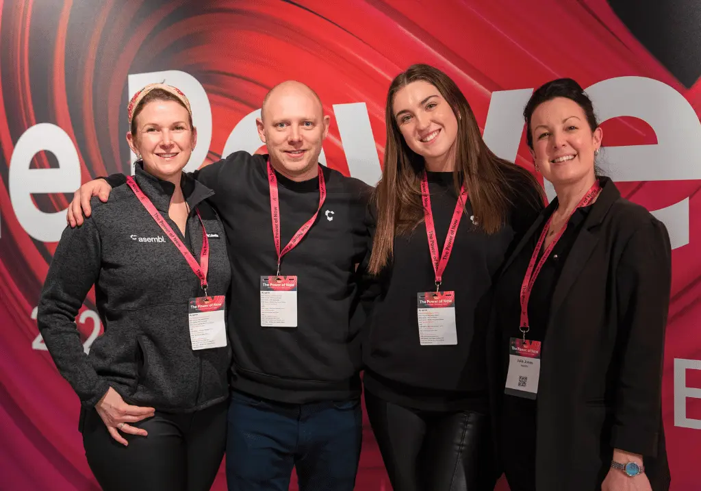 Four asembl event team members standing in front of Dynamic Planner branded background at the conference. They are wearing black asembl uniform