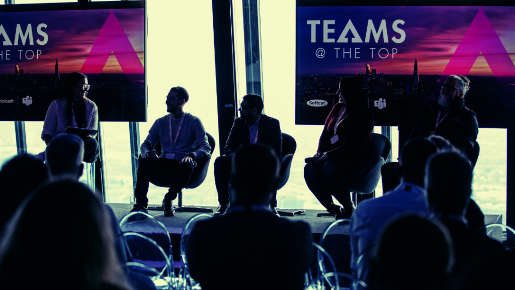 A row of speakers sitting on a stage during a panel session. They are sitting in front of a large window with Teams at the top signage and the silhouette of the audience watches