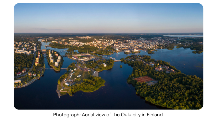 Aerial view of a city surrounded by water and lush greenery, with scattered buildings and dense forests under a clear, blue sky at sunset.